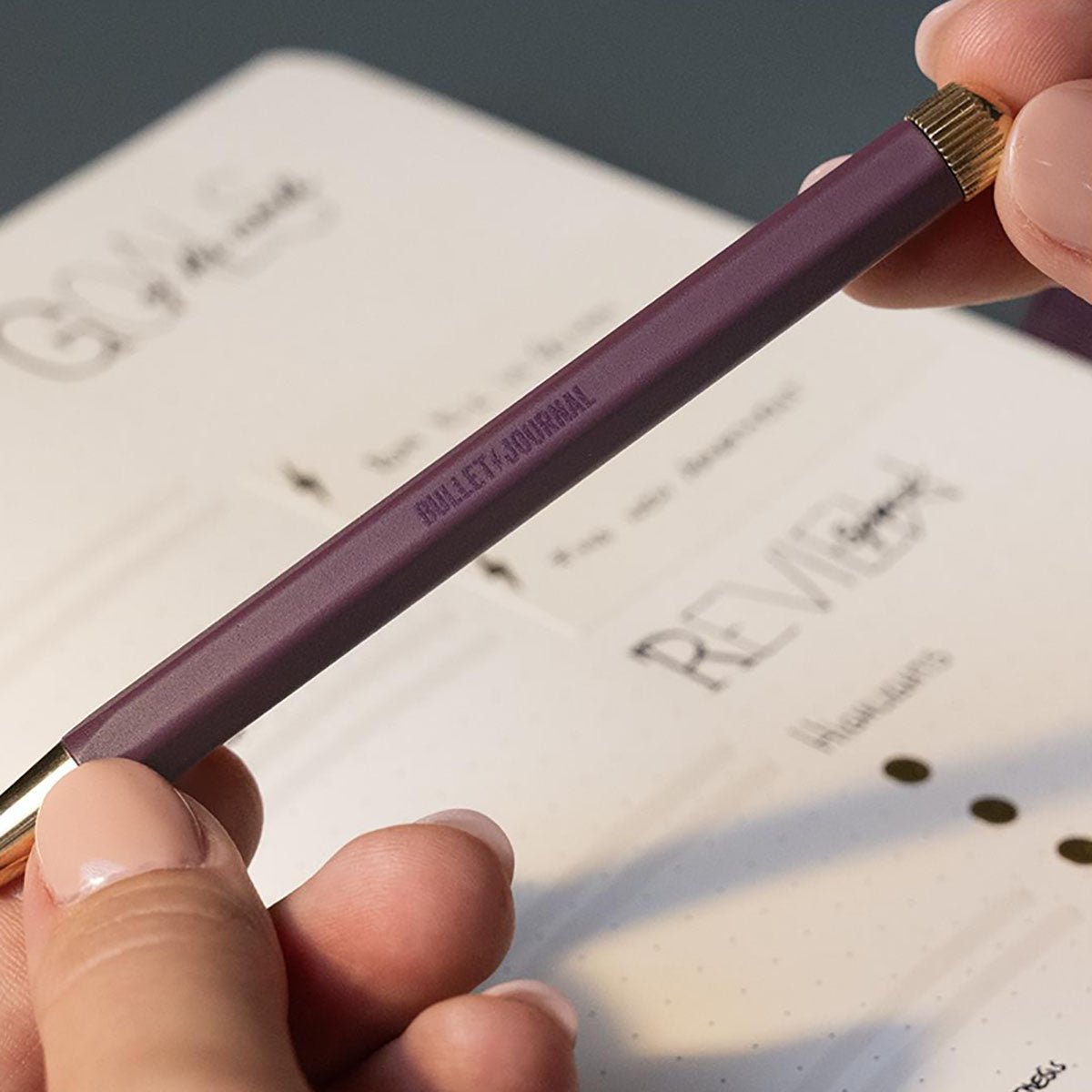 Długopis żelowy z napisem Bullet Journal w kolorze śliwkowym ze złotymi detalami trzymany w dłoniach.